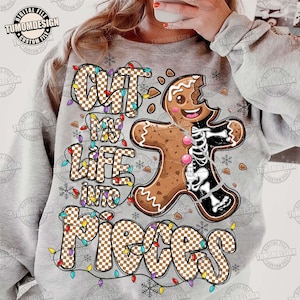 May include: A light gray sweatshirt with the text "Cut My Life Into Pieces" in a checkered pattern, with a gingerbread man graphic. The gingerbread man is half-eaten, revealing a skeleton. Colorful lights and snowflakes decorate the design.