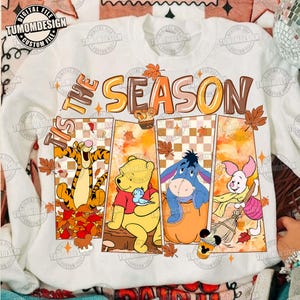 May include: A white long-sleeve sweatshirt featuring the text 'TIS THE SEASON' in orange and brown. The graphic includes four vertical panels displaying Tigger, Winnie the Pooh, Eeyore, and Piglet surrounded by autumn leaves and pumpkins.