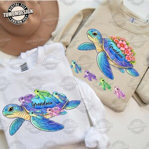 May include: Two sweatshirts, one white and one tan, feature a blue sea turtle graphic with the word "Grandma" and smaller turtles in various colors. The design includes pink flowers and the text "TUMOMDESIGN CUSTOM FILES".