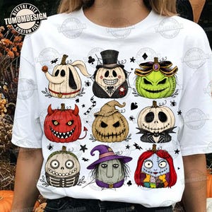 May include: A white t-shirt featuring a graphic print of nine stylized pumpkins decorated as various characters. The design includes small black spiders, stars, and card suit symbols scattered between the pumpkins on the front of the shirt.