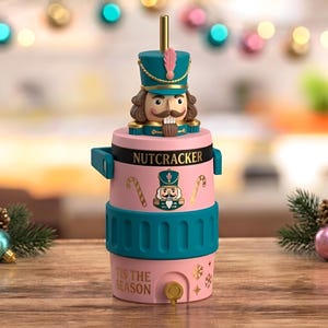 May include: A pink and teal cylindrical beverage container featuring a nutcracker figure on the lid with a gold straw. The body displays the text NUTCRACKER and TIS THE SEASON, decorated with candy cane and snowflake graphics and a small gold spigot.