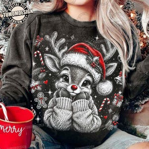 Può includere: Una felpa grigio antracite con la grafica di una renna che indossa un cappello da Babbo Natale rosso e un maglione lavorato a maglia. Il design include elementi festivi come bastoncini di zucchero, un omino di pan di zenzero e bacche.