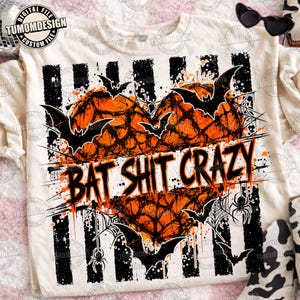May include: A cream-colored t-shirt featuring a graphic of an orange heart with a black spiderweb pattern, black bats, and spiders. The text "BAT SHIT CRAZY" is printed in black across the center. The design includes black vertical stripes.