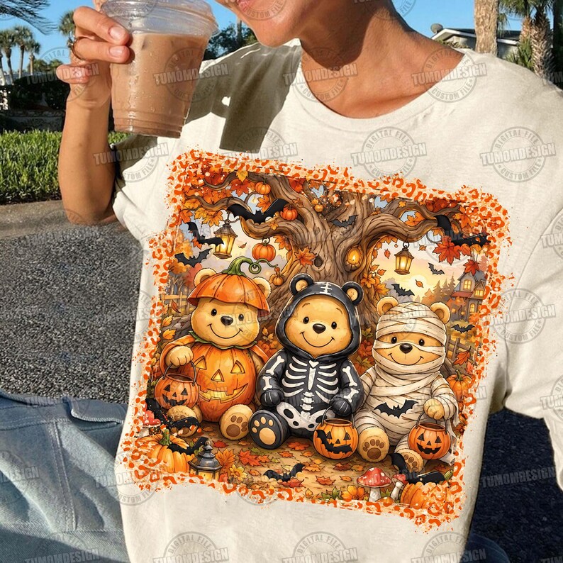 May include: A cream-colored t-shirt featuring a graphic print of three bear characters dressed in Halloween costumes: one as a pumpkin, one as a skeleton, and one as a mummy. The scene includes a large tree, bats, and orange leopard print borders.
