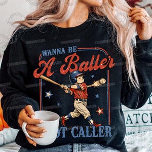 May include: Black sweatshirt with a baseball-themed graphic. The design features a baseball player swinging a bat, with the text "WANNA BE A Baller NOT CALLER" in red and blue. Stars and a baseball complete the design.