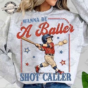 May include: A light gray sweatshirt with a baseball-themed graphic. The graphic features a young baseball player swinging a bat, with the words "WANNA BE A Baller SHOT CALLER" in red and blue. Stars and baseballs are also included.