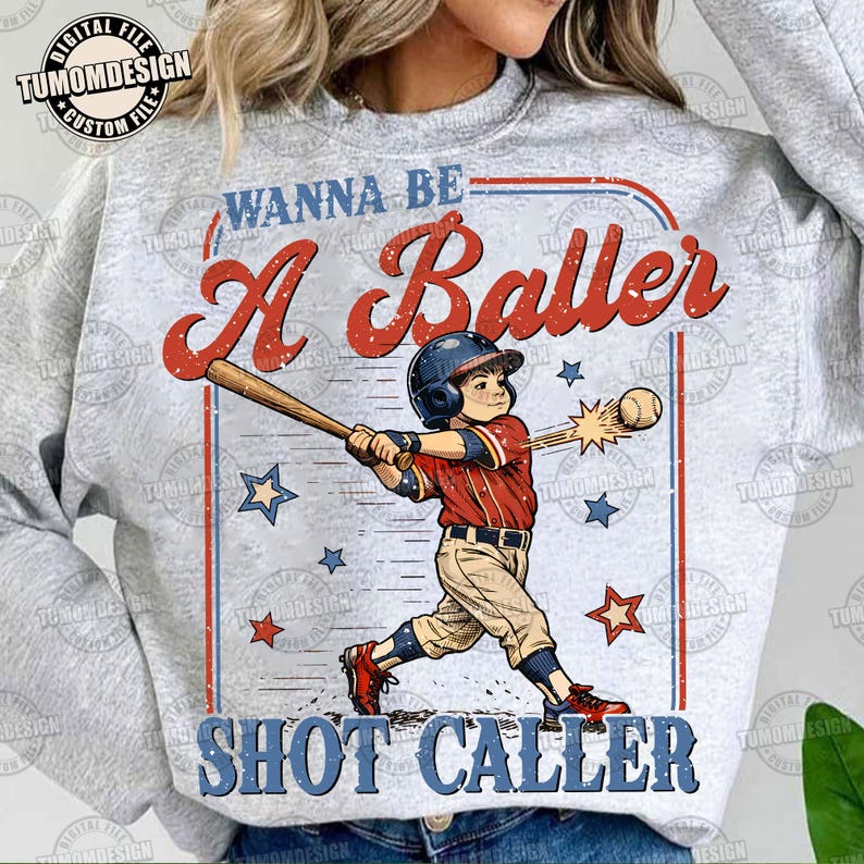 May include: A light gray sweatshirt with a baseball-themed graphic. The graphic features a young baseball player swinging a bat, with the words "WANNA BE A Baller SHOT CALLER" in red and blue. Stars and baseballs are also included.