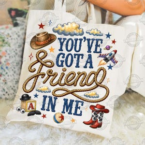 May include: A white canvas tote bag featuring a graphic print with the text "YOU'VE GOT A FRIEND IN ME" in blue letters. The design includes a brown cowboy hat, a coiled rope, a toy block with the letter A, a ball, a rocket, and cowboy boots.