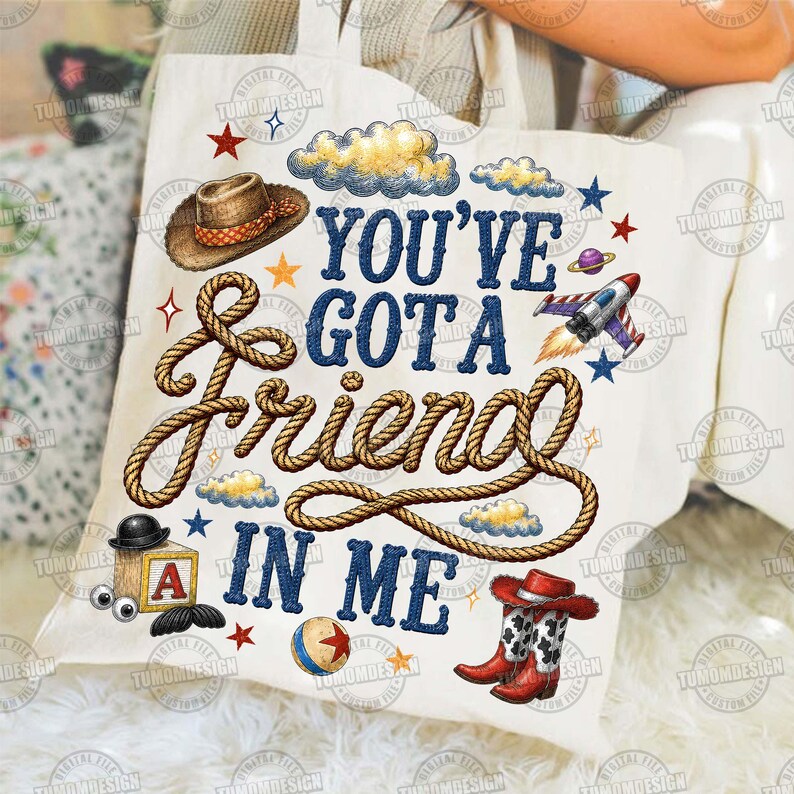 May include: A white canvas tote bag featuring a graphic print with the text "YOU'VE GOT A FRIEND IN ME" in blue letters. The design includes a brown cowboy hat, a coiled rope, a toy block with the letter A, a ball, a rocket, and cowboy boots.