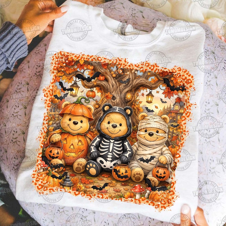 May include: A white sweatshirt featuring a graphic of three teddy bears dressed for Halloween. One bear wears a pumpkin costume, one a skeleton suit, and one is wrapped as a mummy. The scene includes autumn trees, bats, and lanterns.
