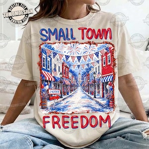 May include: Cream-colored t-shirt featuring a patriotic design with the words "SMALL TOWN FREEDOM" in red and blue. The graphic depicts a street lined with buildings, flags, and fireworks, evoking a festive atmosphere.