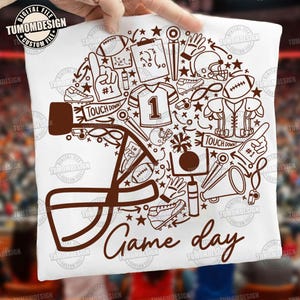 May include: A white t-shirt featuring a brown graphic design of a football helmet silhouette filled with various sports icons including a jersey with the number 1, footballs, a megaphone, a whistle, and the text "TOUCH DOWN" and "Game day".
