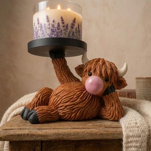 Peut inclure: Une figurine de vache Highland brune allongée soutenant un socle à bougie circulaire noir. La vache a des cornes blanches, des sabots noirs et une bulle rose sur le museau. Une bougie à trois mèches avec un motif floral lavande est posée.