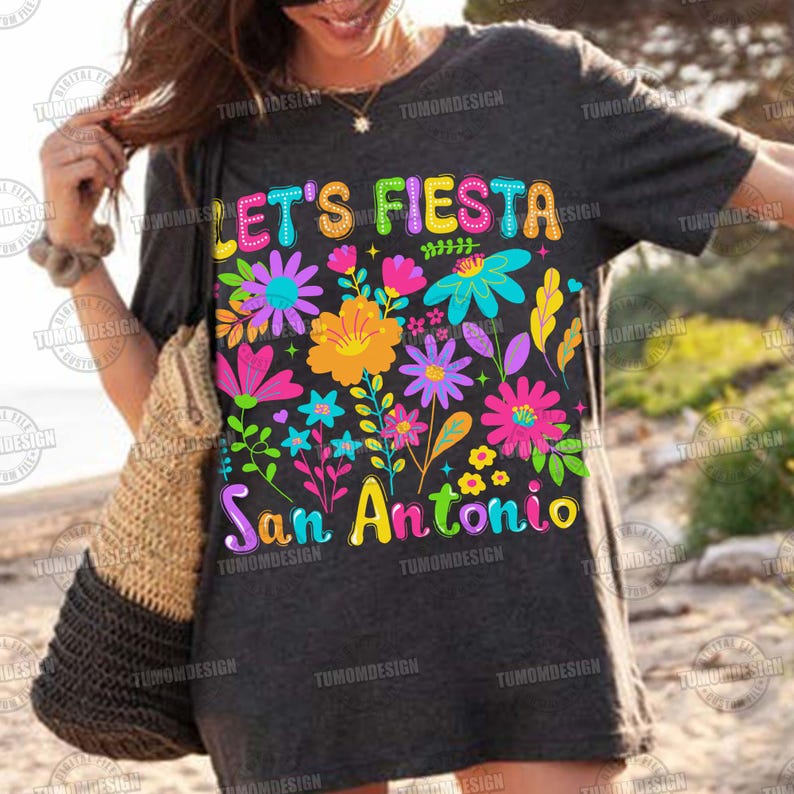 Op de afbeelding: Donkergrijs T-shirt met de tekst "LET'S FIESTA San Antonio" in kleurrijke letters. Het shirt heeft een levendig bloemenontwerp met verschillende bloemen in roze, gele, blauwe en paarse tinten.