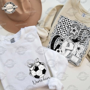 May include: White sweatshirt with a black and white graphic of a soccer ball with a bow on top. The text "Name here" is below the graphic.