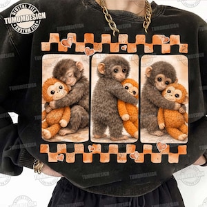 May include: Black sweatshirt featuring a three-panel design of hugging monkeys. The monkeys are gray and orange, with a checkerboard and heart border. The text "TUMOMDESIGN" is visible.