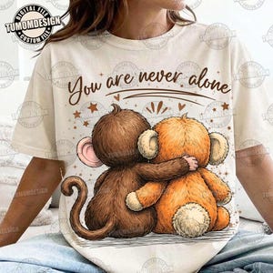 May include: Cream-colored t-shirt featuring a graphic of two hugging plush monkeys. The monkeys are brown and orange, with the text "You are never alone" above them. The design includes stars and hearts.