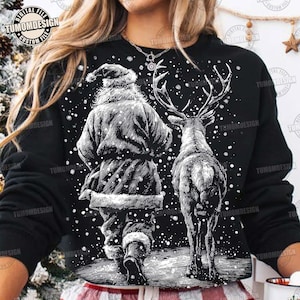 May include: Black sweatshirt featuring a detailed black and white illustration of Santa Claus walking with a reindeer in a snowy scene. The design includes falling snowflakes and the text "TUMOMDESIGN" in a circular logo.