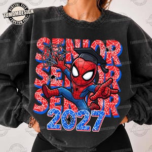 May include: A charcoal black crewneck sweatshirt featuring a graphic of a cartoon superhero wearing a graduation cap. The design includes the text SENIOR in red and blue spiderweb patterns, with the year 2027 printed in large blue digits.