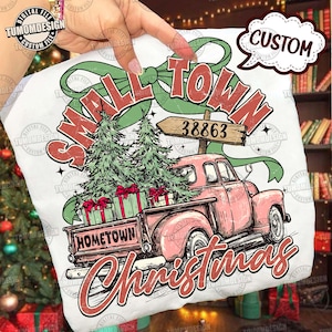 May include: A white t-shirt featuring a pink vintage truck carrying Christmas trees and gifts. The text reads "Small Town Christmas" and "Hometown" on the truck. A sign displays the number 38863.