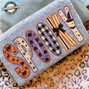 May include: A heather gray sweatshirt featuring the word SPOOKY in large, decorative letters. Each letter has a unique pattern including orange with black cats, cream with spiderwebs, purple with pumpkins, black plaid, orange stripes with a black bow, and cream with a witch hat.