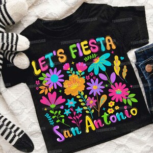 Op de afbeelding: Zwart T-shirt met de tekst "Let's Fiesta San Antonio" in kleurrijke letters. Het shirt heeft een bloemenontwerp met verschillende bloemen in roze, oranje, blauwe en paarse tinten. Het shirt is gemaakt van een zacht materiaal.