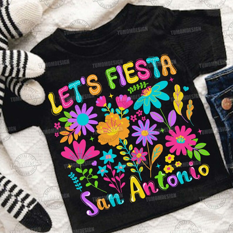 Op de afbeelding: Zwart T-shirt met de tekst "Let's Fiesta San Antonio" in kleurrijke letters. Het shirt heeft een bloemenontwerp met verschillende bloemen in roze, oranje, blauwe en paarse tinten. Het shirt is gemaakt van een zacht materiaal.