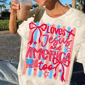 May include: Cream-colored t-shirt with a patriotic design. The shirt features the text "Loves Jesus and America too" in red, with red and blue stripes, bows, stars, and an American flag graphic.
