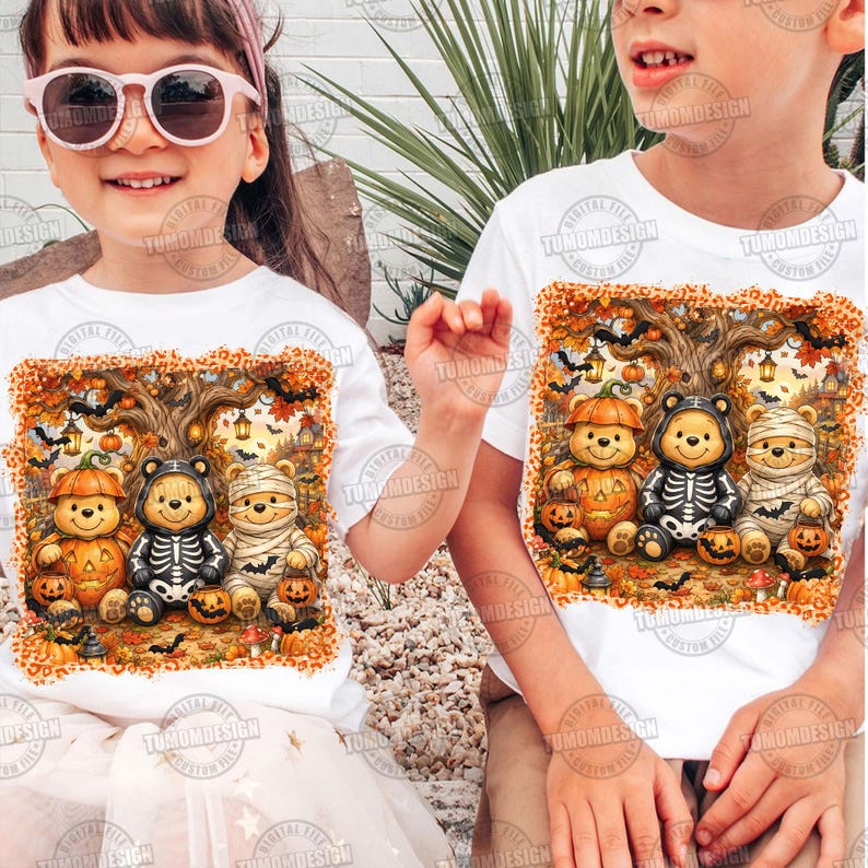 May include: Two white t-shirts featuring a graphic print of three teddy bears dressed in Halloween costumes. One bear wears a pumpkin suit, one a skeleton outfit, and one a mummy wrap. The scene includes a tree, lanterns, and bats.