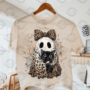 May include: Cream-colored t-shirt featuring a Halloween-themed design. The graphic includes a ghost wearing a leopard print bow and holding a black cat. Additional elements include bats, spiderwebs, and stars. The text "TUMOMDESIGN" is repeated around the image.