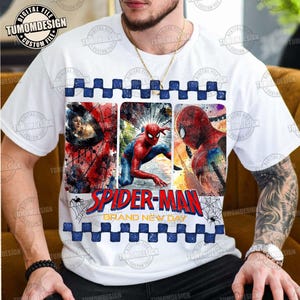 May include: A white short-sleeve t-shirt featuring a graphic print of Spider-Man in three vertical panels. The design includes the text Spider-Man Brand New Day in red and yellow, framed by blue and white checkered borders and spider webs.