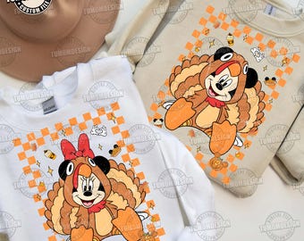 Può includere: Due felpe, una bianca e una beige, presentano personaggi dei cartoni animati vestiti da tacchini. I personaggi sono ambientati su uno sfondo a scacchi arancione e bianco con grafica a tema autunnale. Il testo "TUMOMDESIGN" è visibile.