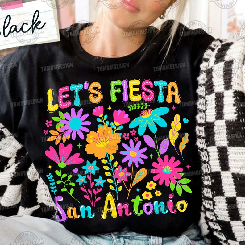 Op de afbeelding: Zwart T-shirt met de kleurrijke tekst "LET'S FIESTA San Antonio". Het shirt heeft een levendig bloemenontwerp met bloemen in roze, oranje, blauwe en paarse tinten.