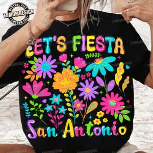 Op de afbeelding: Zwart T-shirt met een kleurrijk bloemmotief en de tekst "LET'S FIESTA San Antonio" in heldere, speelse letters. Het ontwerp toont diverse bloemen in roze, oranje, blauw en paars.