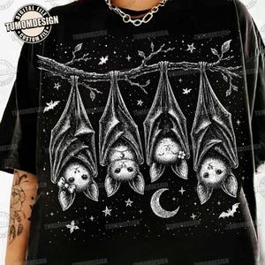 May include: A black t-shirt featuring a white graphic illustration of four bats hanging upside down from a tree branch. The design includes a crescent moon, scattered stars, and small leaves. One bat wears a bow, and another has a small flower.