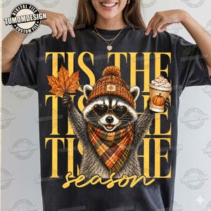 May include: A charcoal gray t-shirt featuring a graphic of a raccoon wearing a brown knit hat, heart-shaped sunglasses, and a plaid scarf. The raccoon holds an orange maple leaf and a coffee cup. The text reads TIS THE SEASON in yellow.