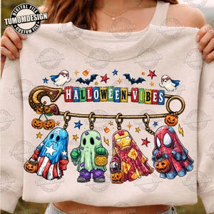 May include: A beige sweatshirt featuring a large safety pin graphic with the text HALLOWEEN VIBES. Hanging from the pin are four ghost figures dressed as superheroes, each holding a small jack-o'-lantern bucket, surrounded by bats and stars.