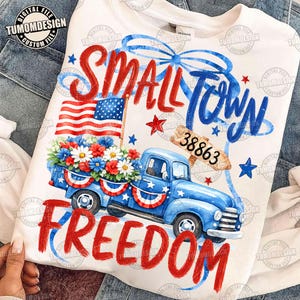 May include: White sweatshirt with a patriotic design. Features a blue pickup truck filled with flowers, an American flag, and the words "Small Town Freedom" in red and blue. Includes the number "38863".