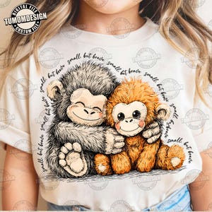 May include: A cream t-shirt with a graphic of two cartoon monkeys embracing. One monkey is grey, the other is orange. The text "small, but brave" is written around the monkeys. The shirt also features the text "TUMOMDESIGN" in the upper left corner.