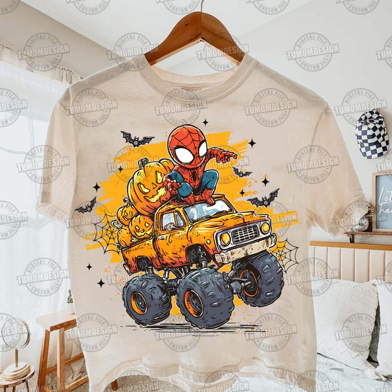May include: A beige t-shirt featuring a cartoon illustration of Spider-Man on a monster truck loaded with pumpkins. The truck is orange, and the background includes bats and a spider web, perfect for Halloween.