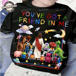 Può includere: Una t-shirt nera con una grafica colorata che mostra una figura centrale in tunica bianca circondata da vari personaggi dei cartoni animati, tra cui un cowboy, un ranger spaziale e un dinosauro verde, con il testo YOU'VE GOT A FRIEND IN ME.