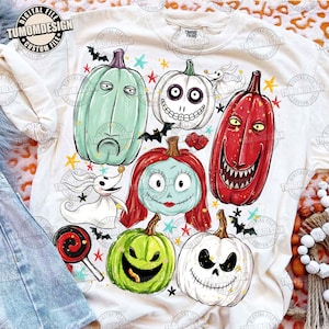 May include: A cream-colored t-shirt featuring a graphic print of various pumpkins with faces, a ghost dog, and small bats. The design includes a tall green pumpkin, a white pumpkin with a skeleton face, a red pumpkin, and a green pumpkin with a wide grin.