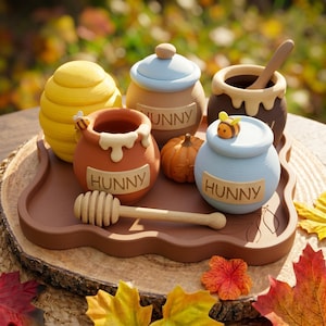 May include: A collection of four small ceramic honey pots in yellow, orange, brown, and light blue, each labeled HUNNY. The set includes a wooden honey dipper, a small orange pumpkin, and two bee figurines, all arranged on a brown tray.