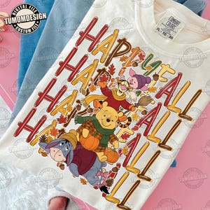 May include: A cream-colored t-shirt featuring a graphic of Winnie the Pooh, Tigger, Piglet, and Eeyore dressed in autumn clothing with pumpkins and leaves. The text "HAPPY FALL Y'ALL" is printed in large, stylized red and orange letters.
