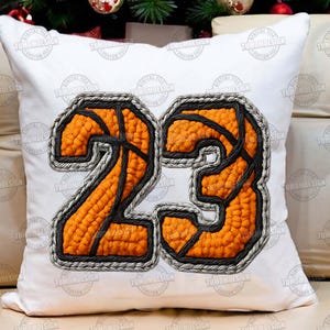 May include: White square cushion displaying the number 23 in an orange basketball design with black lines and a grey border. The cushion has the text "TUMOMDESIGN" repeated around the edges.