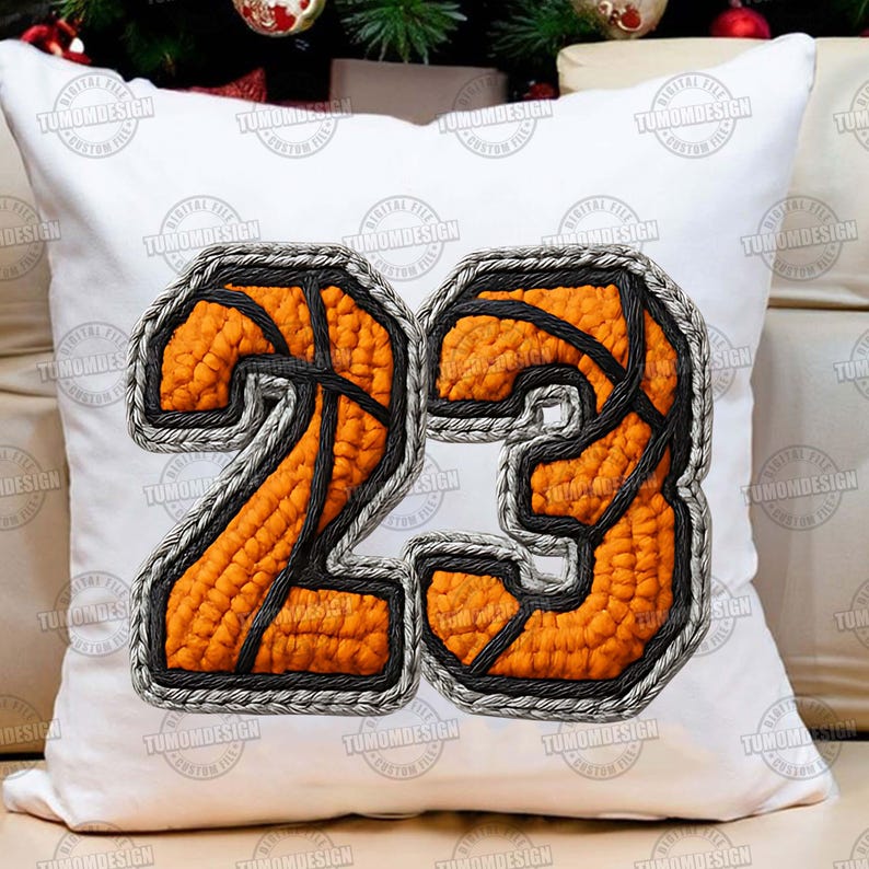 May include: White square cushion displaying the number 23 in an orange basketball design with black lines and a grey border. The cushion has the text "TUMOMDESIGN" repeated around the edges.