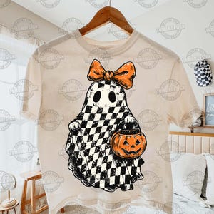 K&ouml;nnte beinhalten: Beiges T-Shirt mit einem Halloween-Geister-Design. Der Geist ist schwarz-wei&szlig; kariert, mit einer orangefarbenen Schleife und einem K&uuml;rbis-Eimer. Das T-Shirt h&auml;ngt an einem Holzb&uuml;gel.