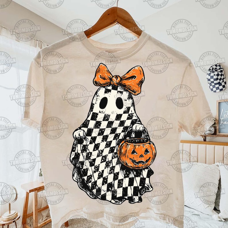 K&ouml;nnte beinhalten: Beiges T-Shirt mit einem Halloween-Geister-Design. Der Geist ist schwarz-wei&szlig; kariert, mit einer orangefarbenen Schleife und einem K&uuml;rbis-Eimer. Das T-Shirt h&auml;ngt an einem Holzb&uuml;gel.