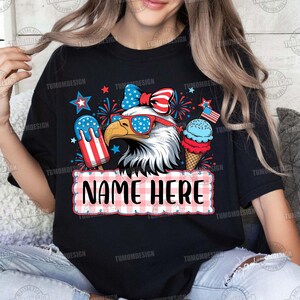 May include: Black t-shirt featuring a patriotic design with an eagle wearing sunglasses, a bow, and ice cream. The design includes the text "NAME HERE" in a red and white checkered frame, and stars.