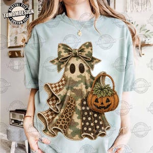May include: A light blue t-shirt featuring a central graphic of a ghost wearing a large bow. The ghost is patterned with camouflage, leopard print, and polka dots, and it is holding a textured orange pumpkin basket with green leaves on top.